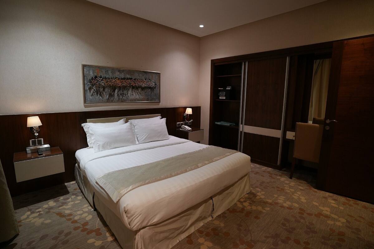 A hotel room with a large bed, a nightstand with a lamp, a wardrobe, and a painting on the wall.