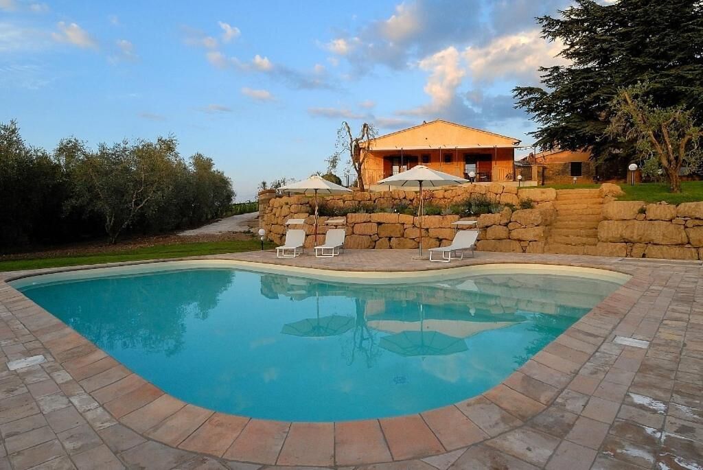 A swimming pool with lounge chairs and umbrellas, surrounded by stone walls and a building with a sloped roof.