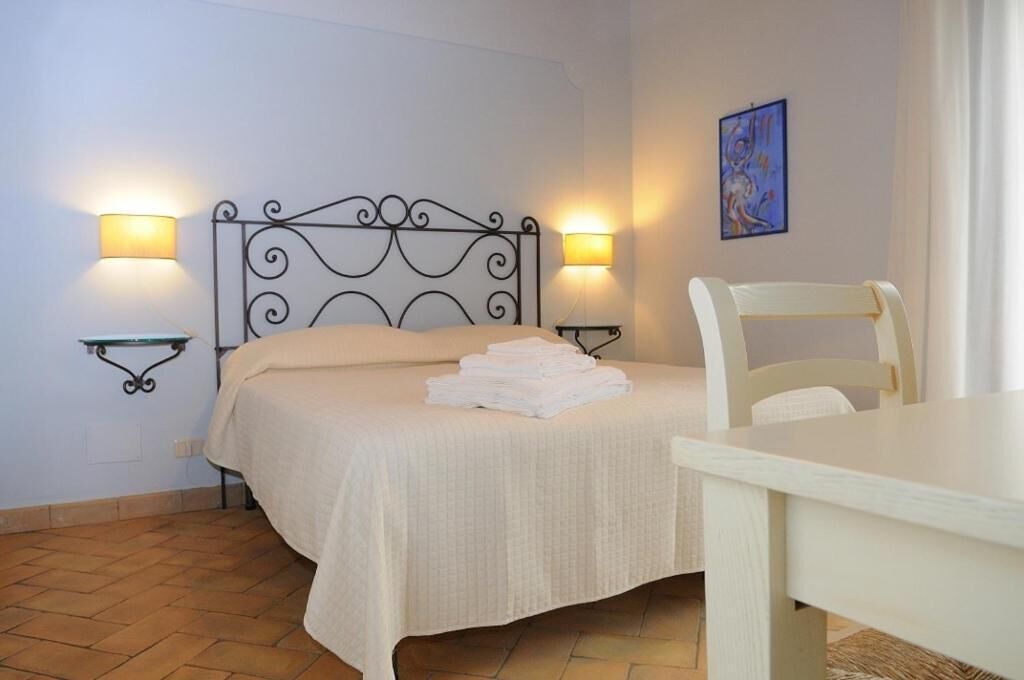 A neatly made bed with a white bedspread and a stack of white towels, two wall-mounted lamps, a painting on the wall, and a wooden table.