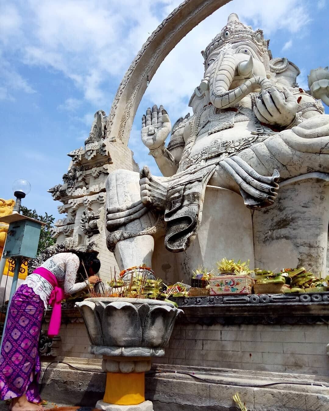 A large stone statue of a deity with intricate carvings, a large bowl in front, and a person performing a ritual.