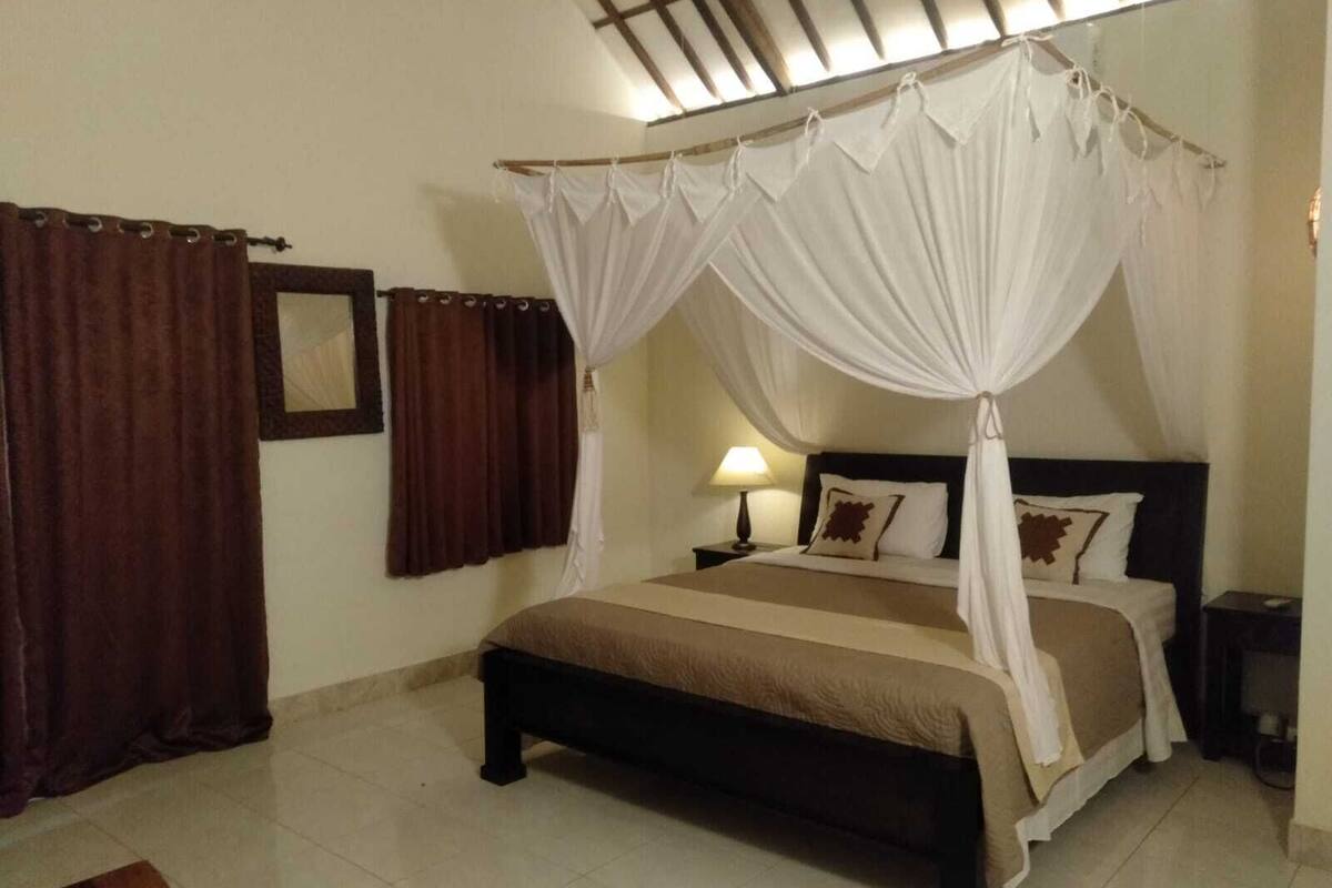 Ein Schlafzimmer mit einem Bett, das von einem Baldachin überdacht wird, einem Nachttisch mit Lampe und einem braunen Teppich auf dem Boden.