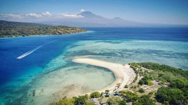 Vista aérea de una zona costera con mar azul cristalino, una playa arenosa y montañas al fondo.