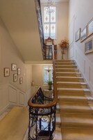 Una gran escalera con barandilla de madera y balaustrada, que conduce a un rellano con un candelabro y cuadros en las paredes.