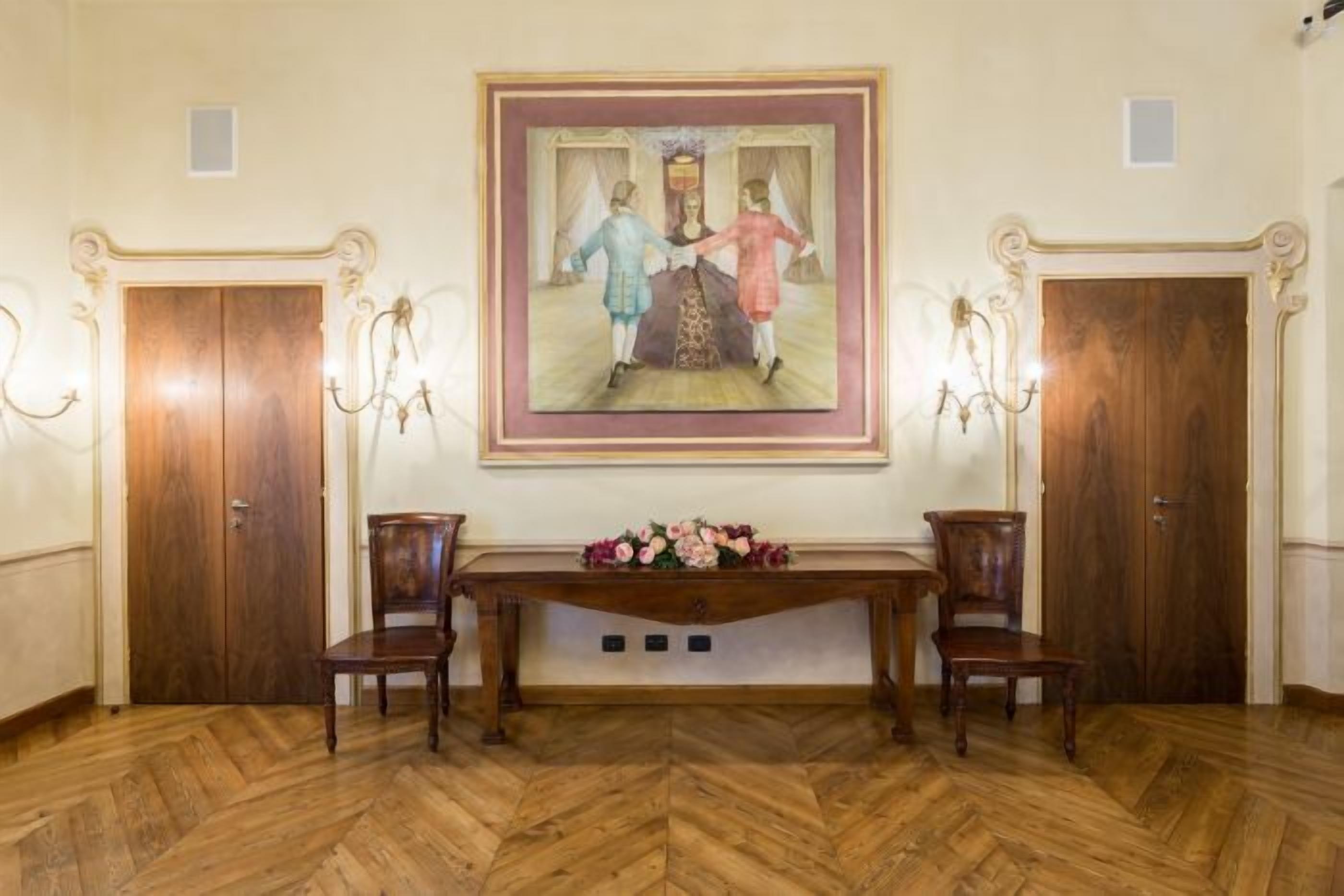 Una habitación con piso de madera en dibujo de espiga, dos sillas de madera, una mesa de madera con un arreglo floral y un cuadro enmarcado sobre la mesa.