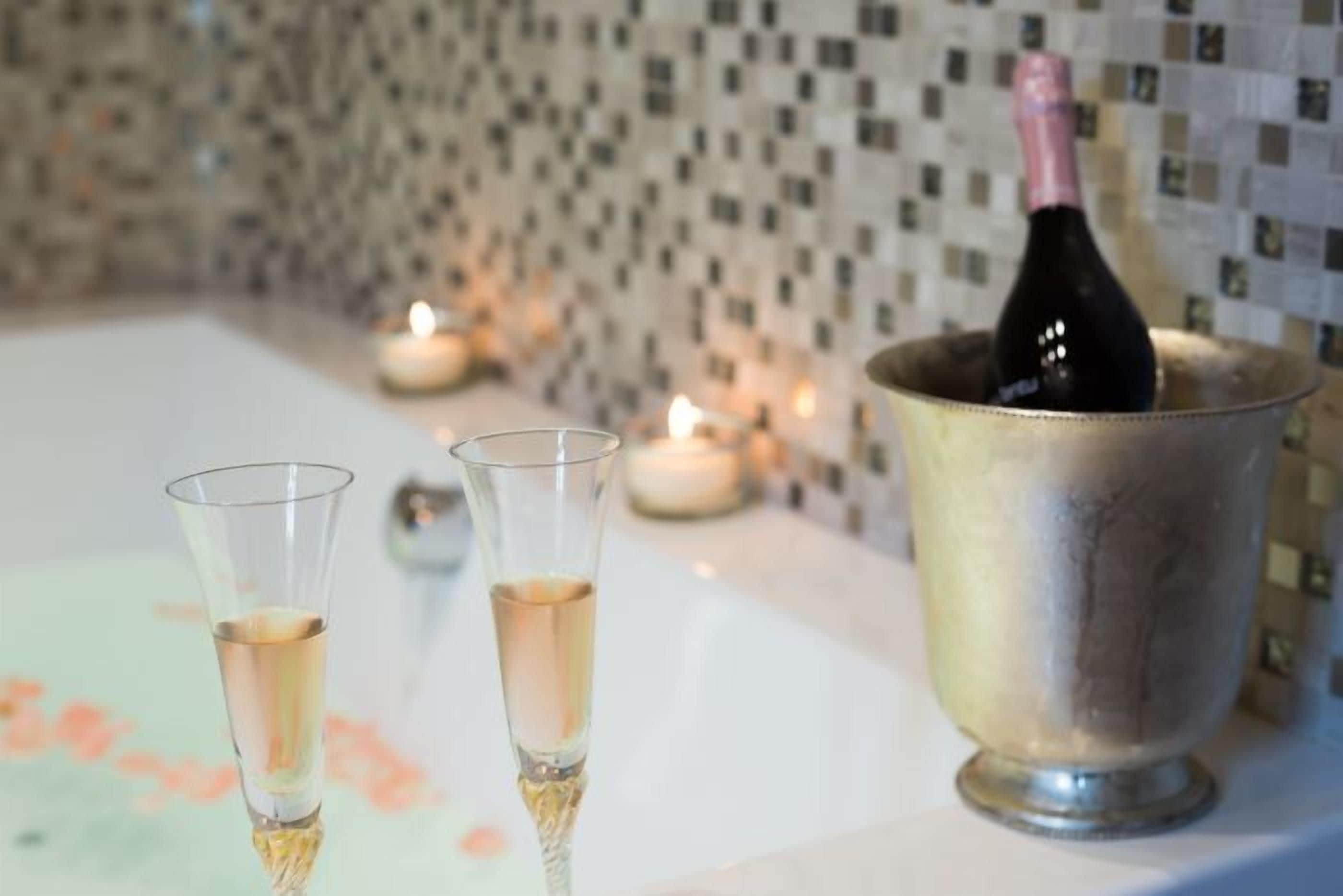 Une bouteille de champagne dans un seau en métal, accompagnée de deux verres remplis de champagne, le tout placé devant un mur carrelé et éclairé par des bougies.