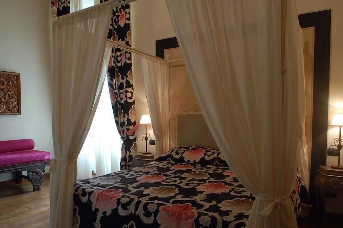 Una cama con dosel, colcha estampada, mesita de noche con lámpara y un banco rosa en una habitación de hotel.