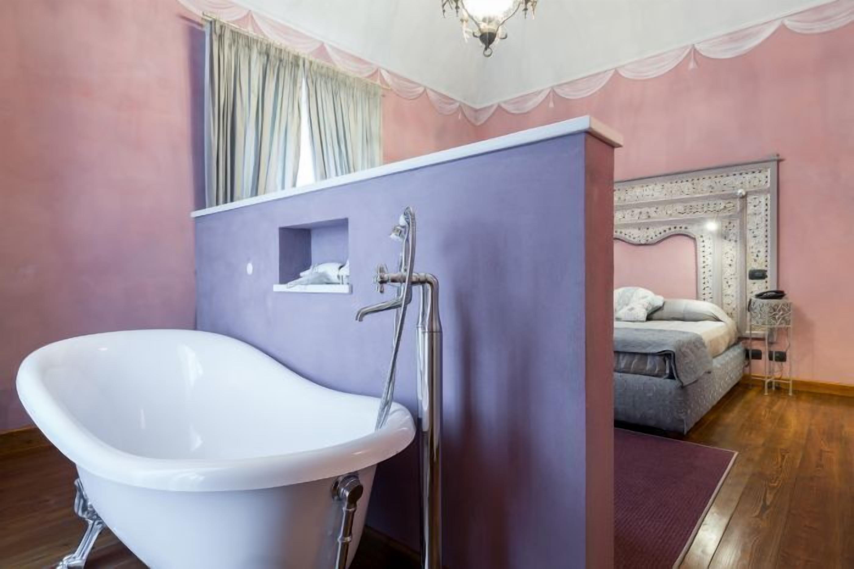 Une salle de bain avec une baignoire sur pieds, un meuble-lavabo avec un miroir, et une chambre adjacente avec un lit agrémenté d’une tête de lit décorative.