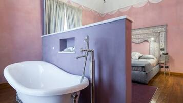 Une salle de bain avec une baignoire sur pieds, un meuble-lavabo avec un miroir, et une chambre adjacente avec un lit agrémenté d’une tête de lit décorative.