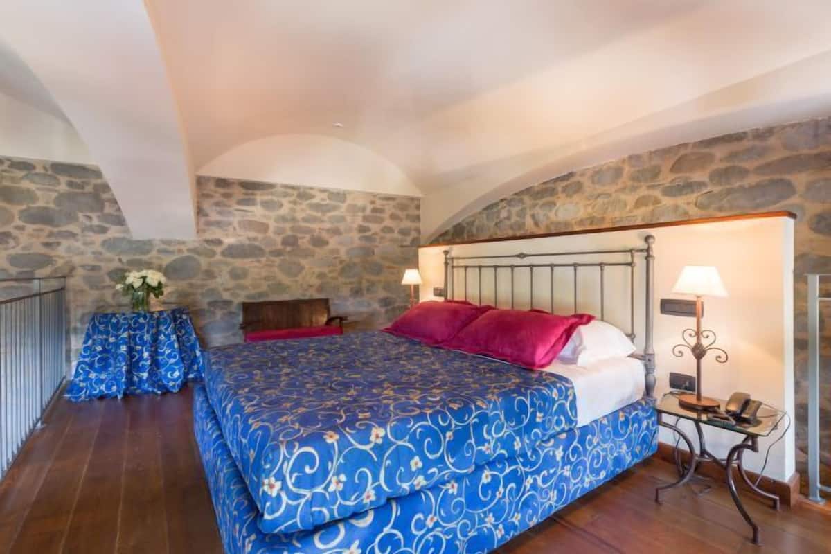 Un dormitorio con pared de piedra, una cama con ropa de cama azul y blanca, dos lámparas de noche y una pequeña mesa con un florero.