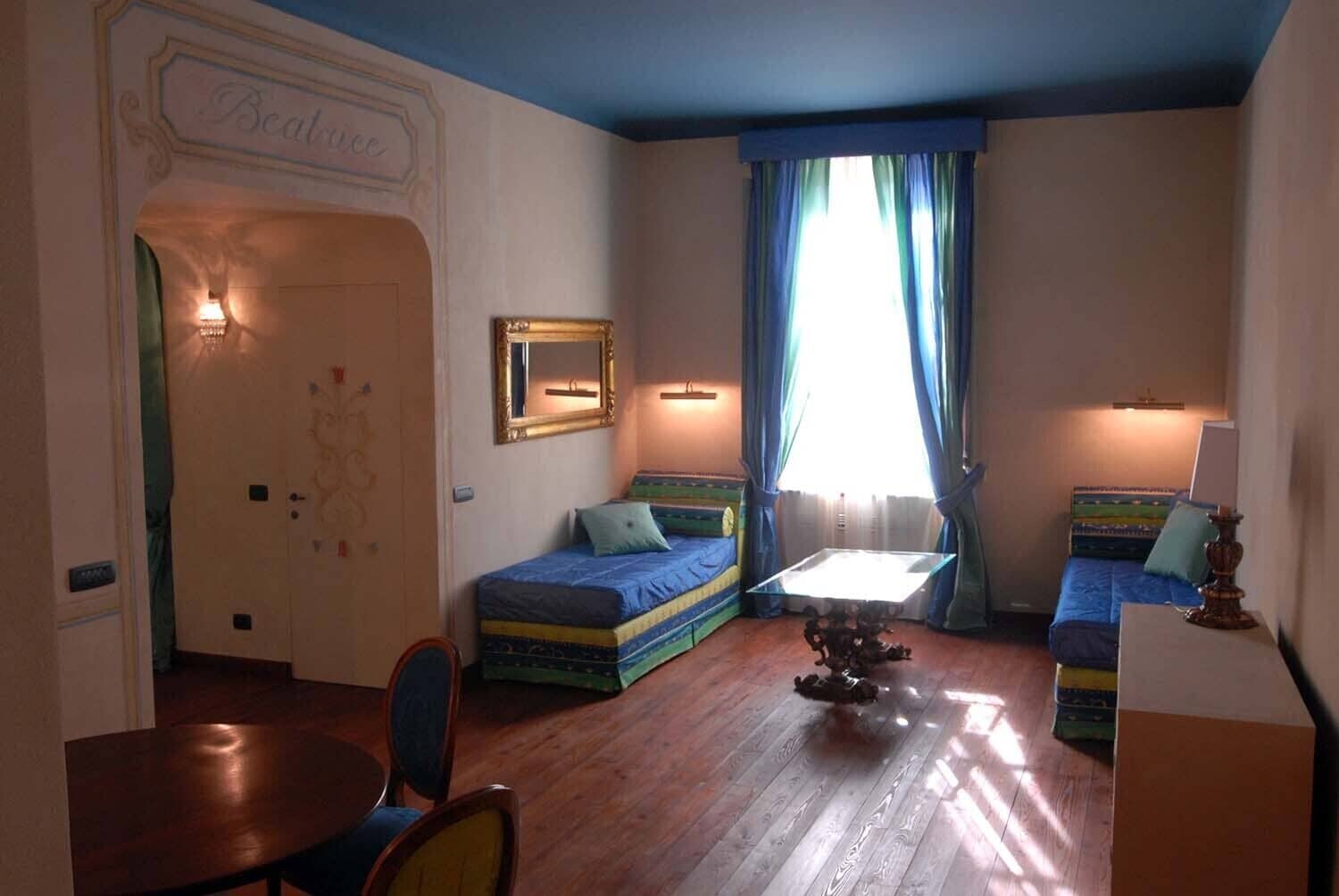 A room with two beds, a small table, and a chair. There is a door labeled "Beatrice" and a mirror on the wall.