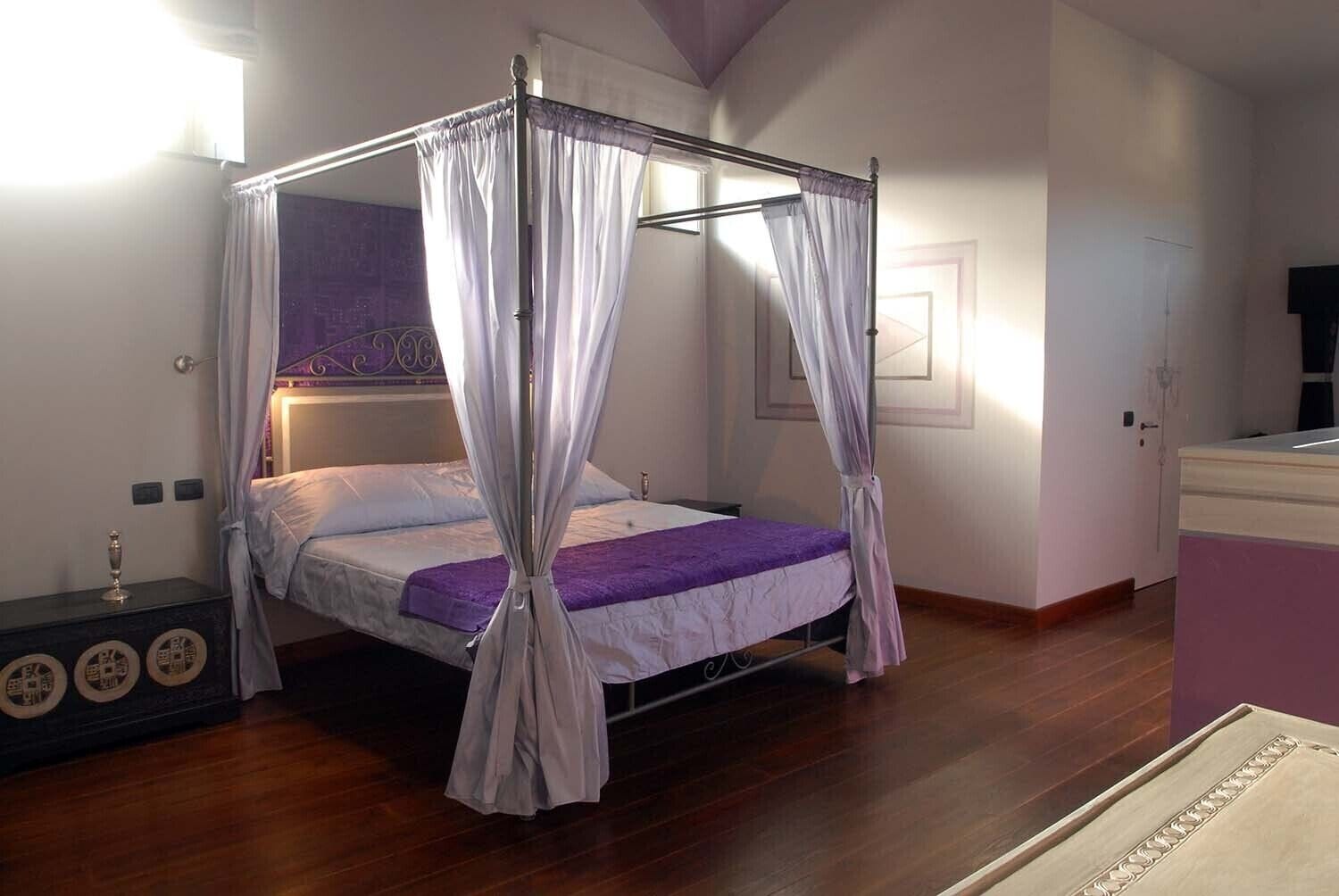 A four-poster canopy bed with a purple bedspread, a bedside table with decorative plates, and a nightstand with a lamp.