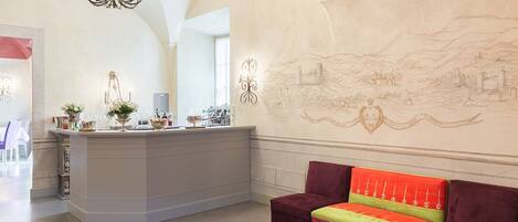 A hotel lobby with a curved reception desk, a chandelier, a sofa with a colorful cushion, and a wall mural.