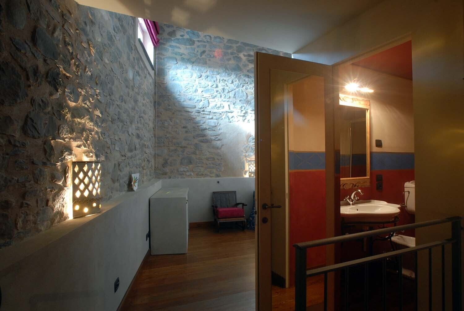 Una habitación con pared de piedra, un refrigerador pequeño, un banco y un baño con lavabo y espejo.