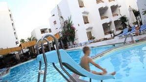 A swimming pool with a metal ladder, surrounded by white buildings with balconies, and a few lounge chairs.