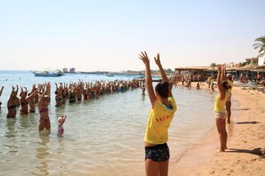 Cena de praia com pessoas na água e na areia, barcos ao fundo e céu claro.