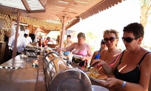 A covered outdoor dining area with metal tables and chairs, people eating, and a sunny environment. - Marlin Inn Azur Resort (Hurghada)
