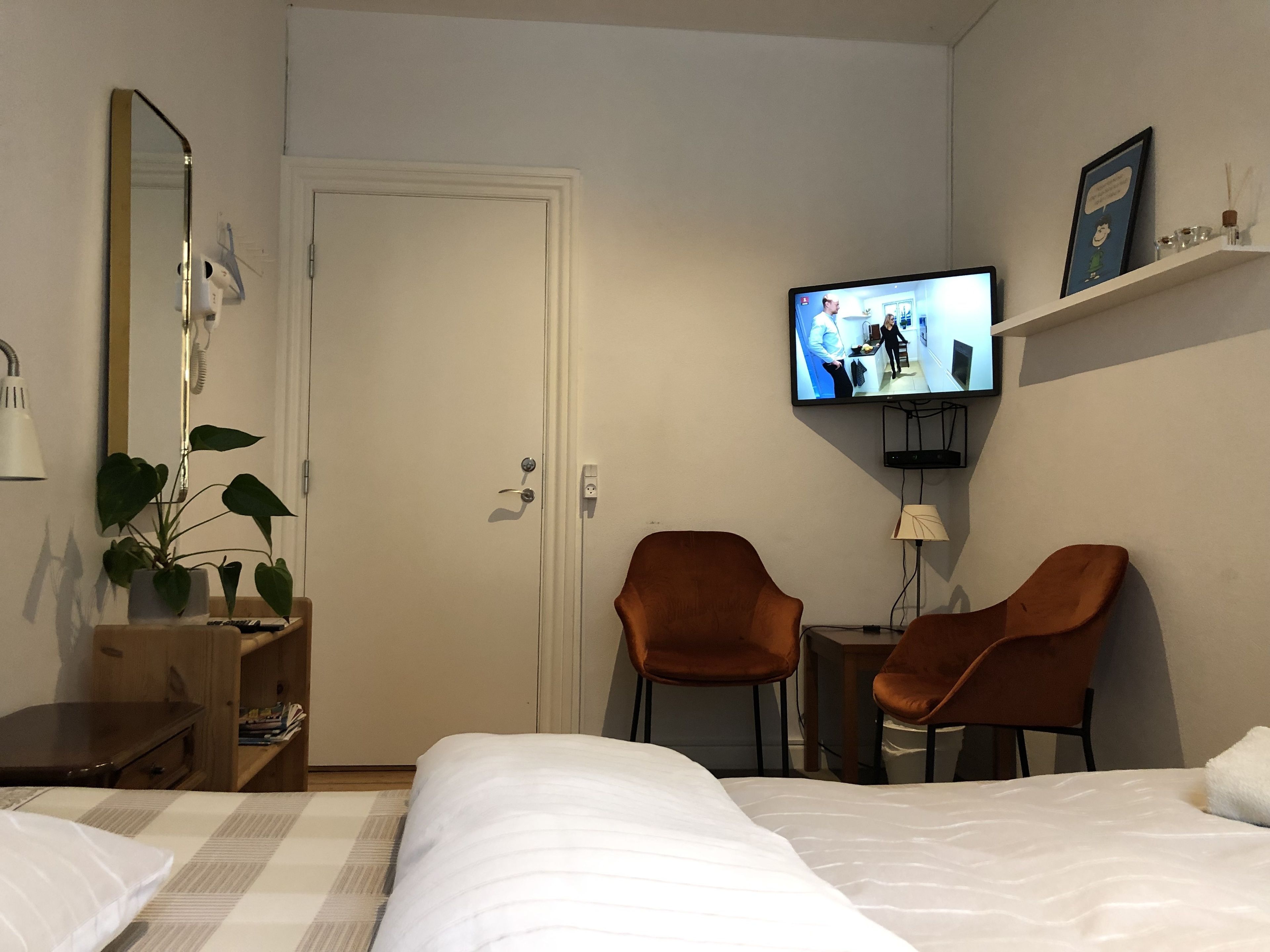 Een hotelkamer met een bed, twee bruine stoelen, een televisie aan de muur, een spiegel, een plant en een kleine tafel met een lamp.
