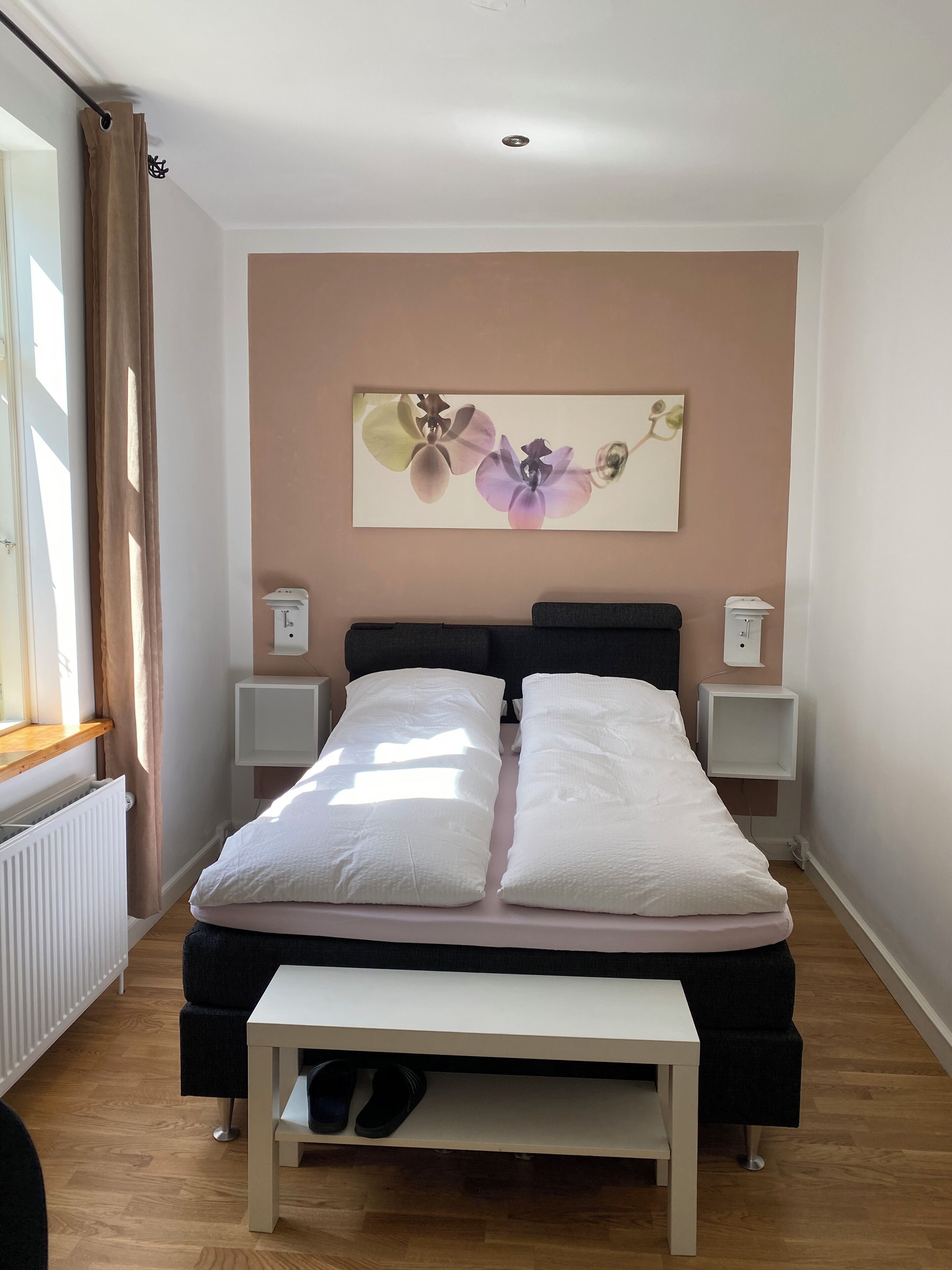 A bedroom with a bed, bedside tables, a nightstand, a window with curtains, a radiator, and a wall-mounted artwork.