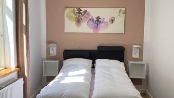 A bedroom with a bed, bedside tables, a nightstand, a window with curtains, a radiator, and a wall-mounted artwork.