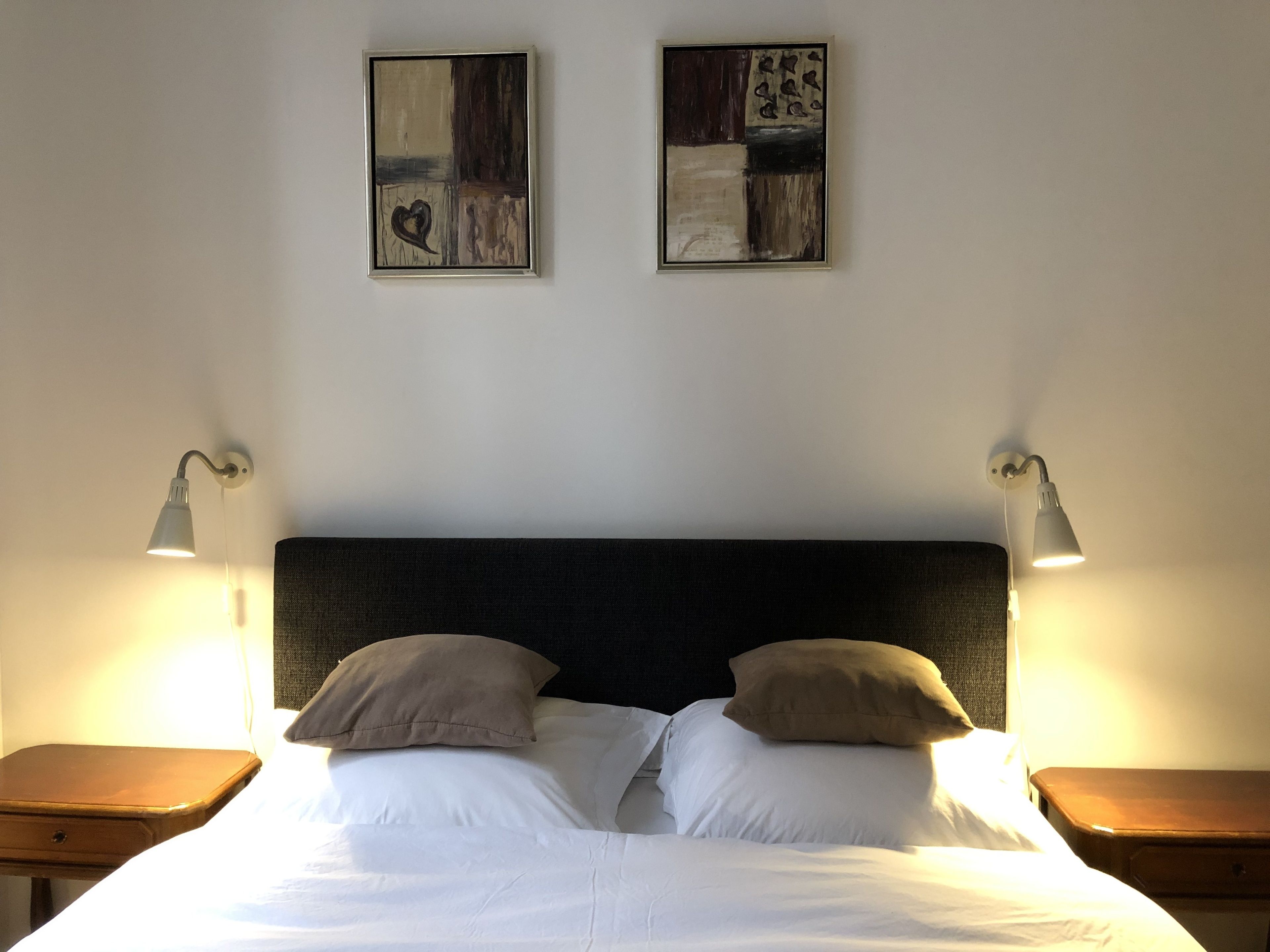 Una cama bien tendida con dos almohadas, dos lámparas fijadas a la pared y dos cuadros abstractos.