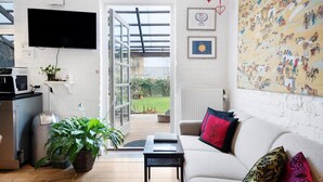A modern living room with a sofa, a coffee table, and a TV mounted on the wall. There is a large window with a view of the garden. - Davaa’S Bed and Breakfast (Randers)