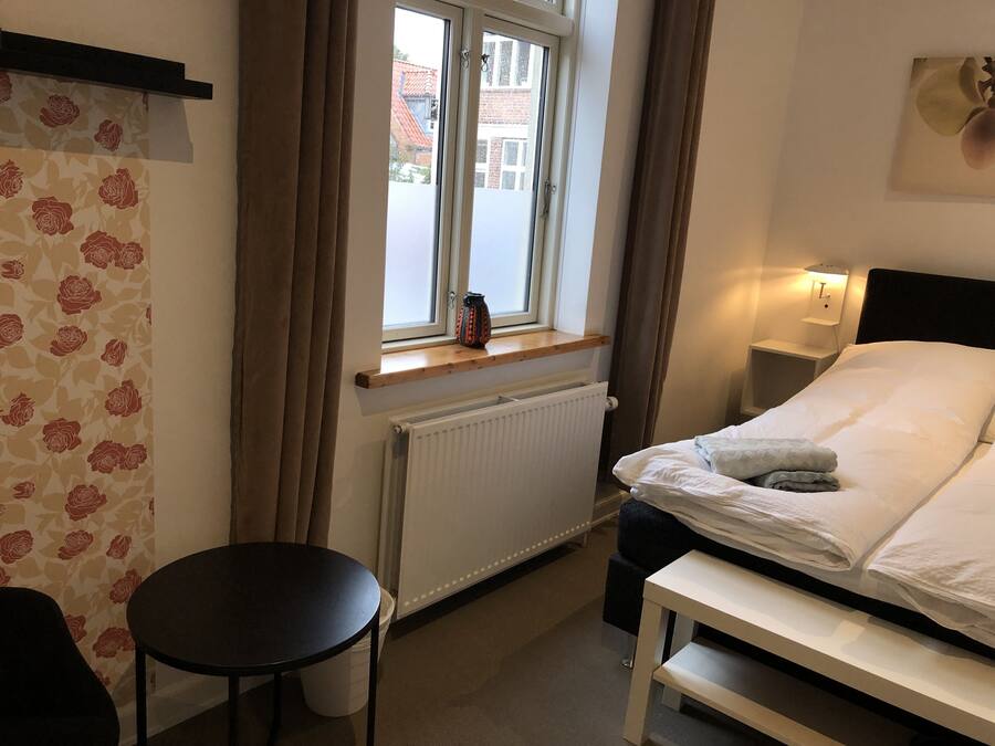 A hotel room with a bed, bedside table, window, radiator, and a small table.