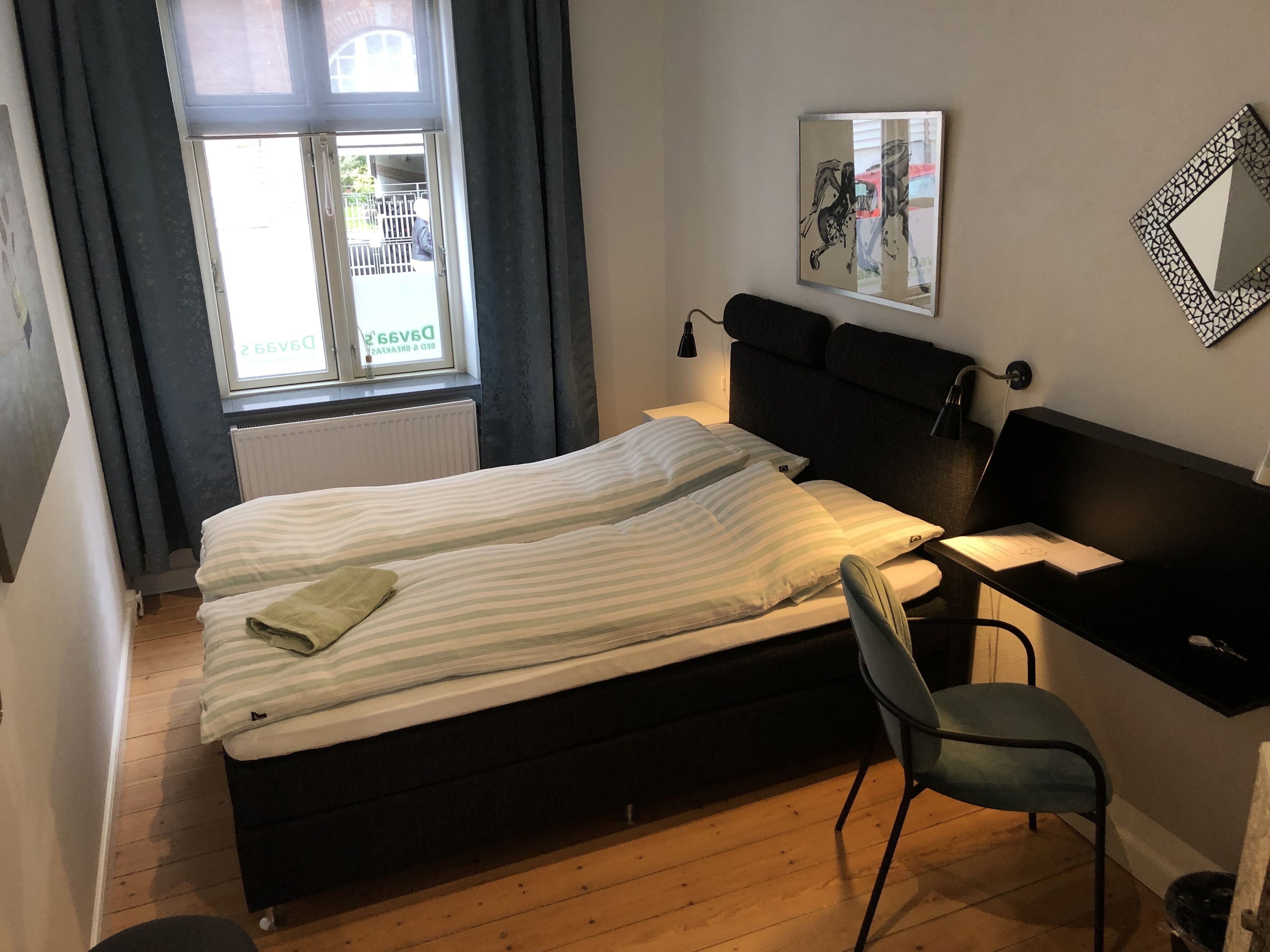 Una camera da letto con un letto, una scrivania, una sedia, un termosifone, una finestra con tende e uno specchio fissato al muro.