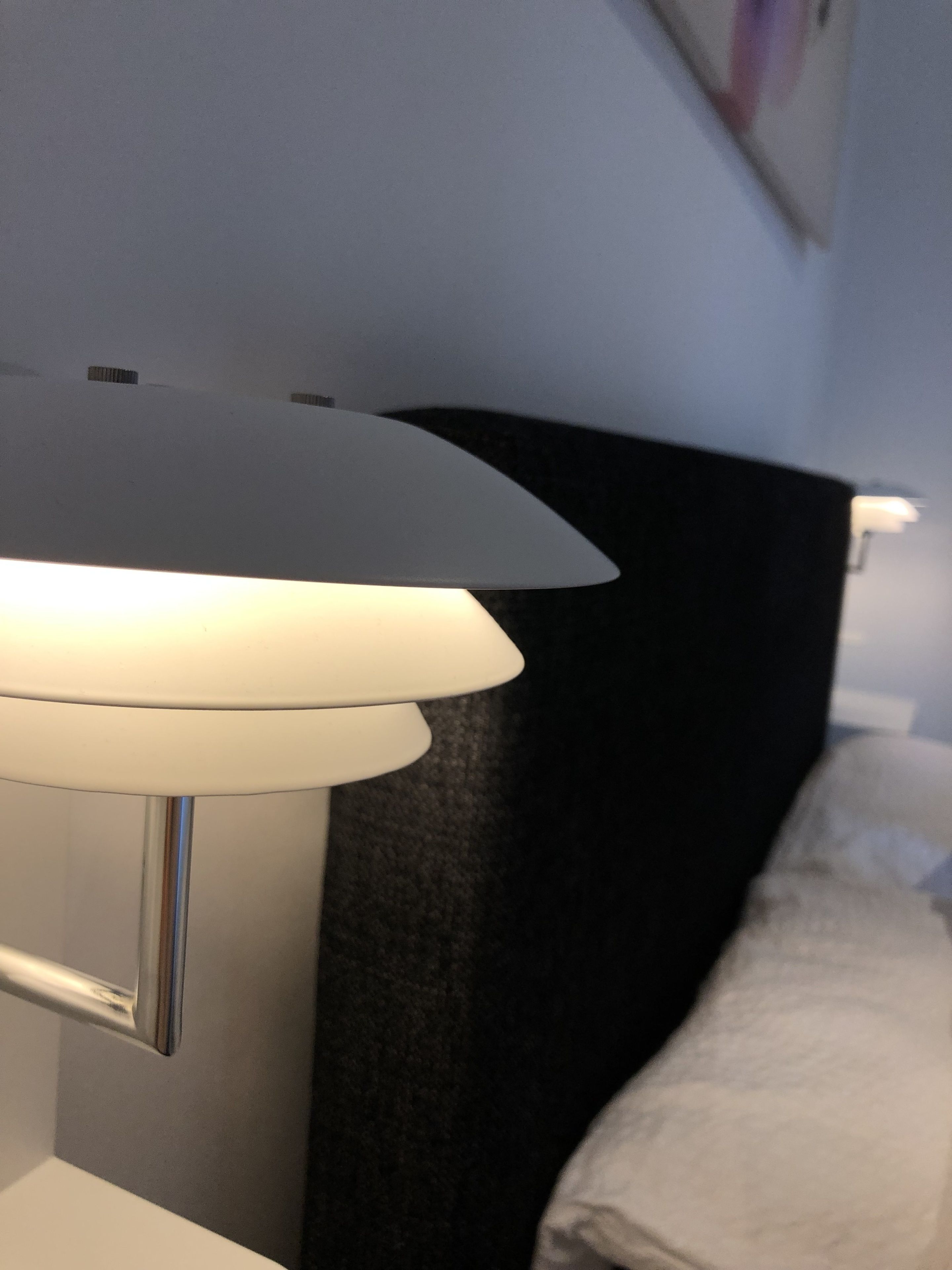 A bedside lamp with a white shade and a metallic base, positioned next to a dark-colored headboard and a white pillow.