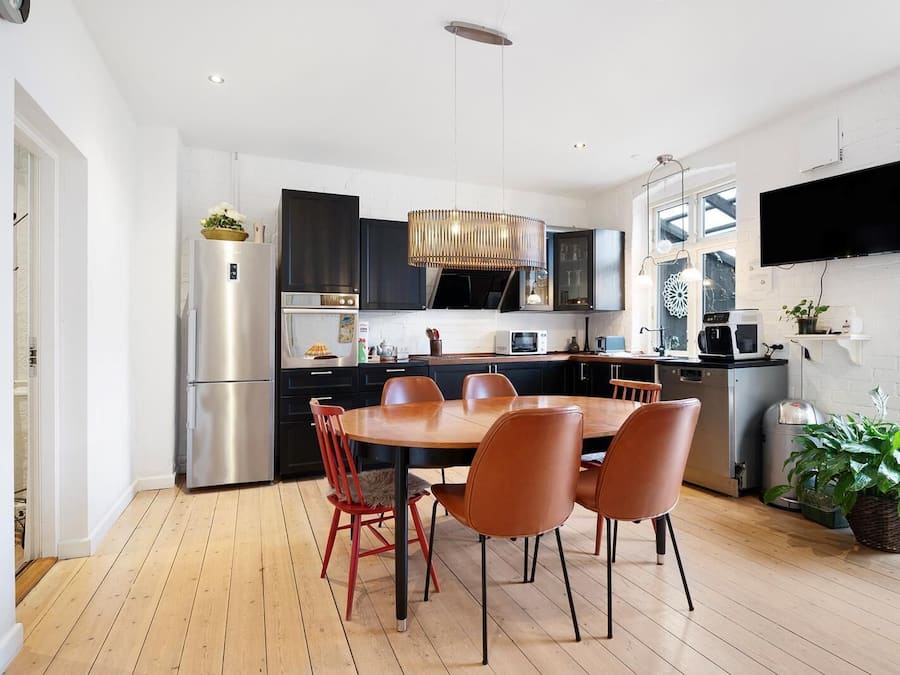 A modern kitchen with a dining area, featuring a large wooden table, red chairs, and stainless steel appliances.