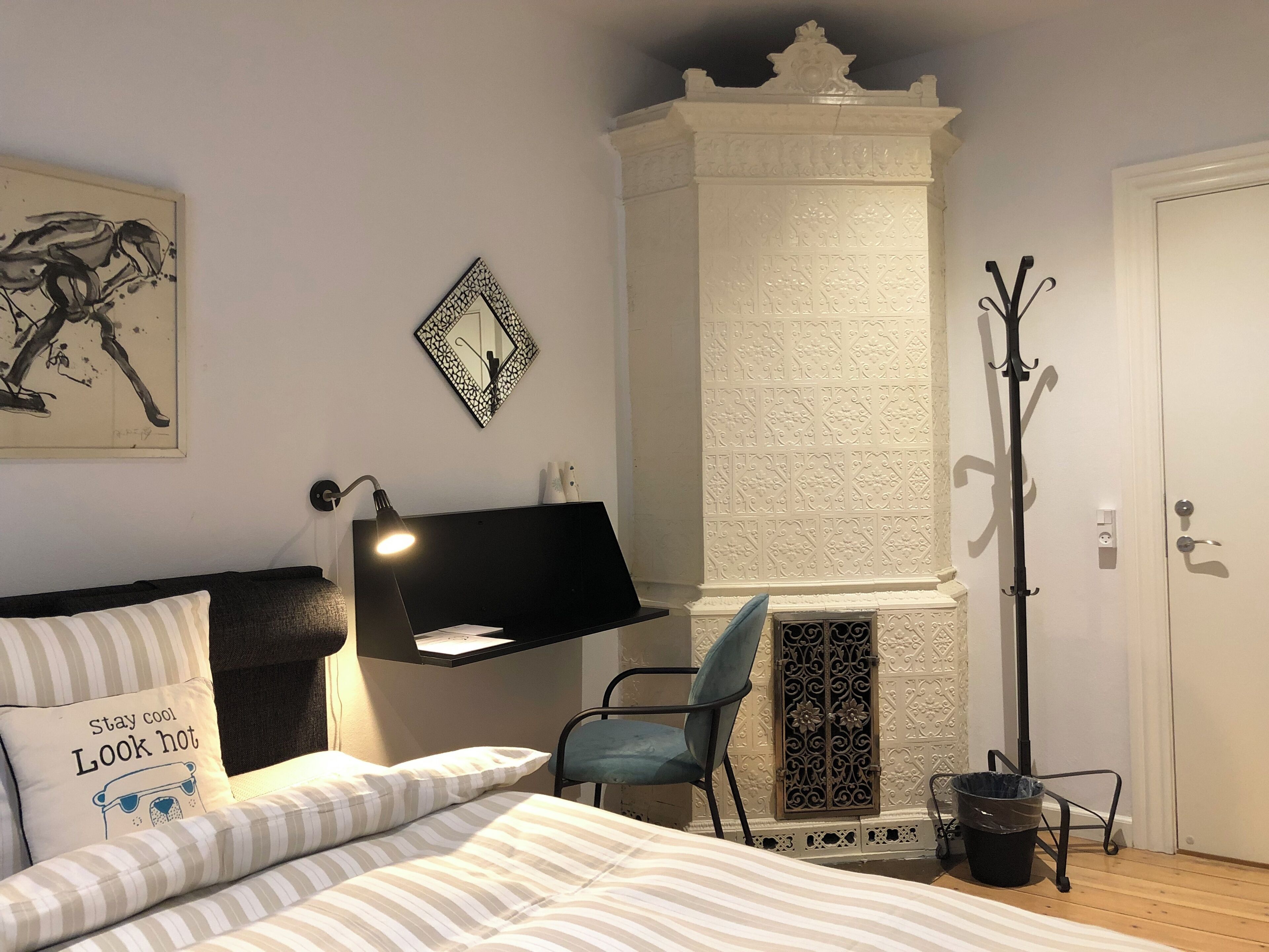 A bedroom with a bed, a desk with a lamp, a chair, a decorative mirror, and a fireplace.