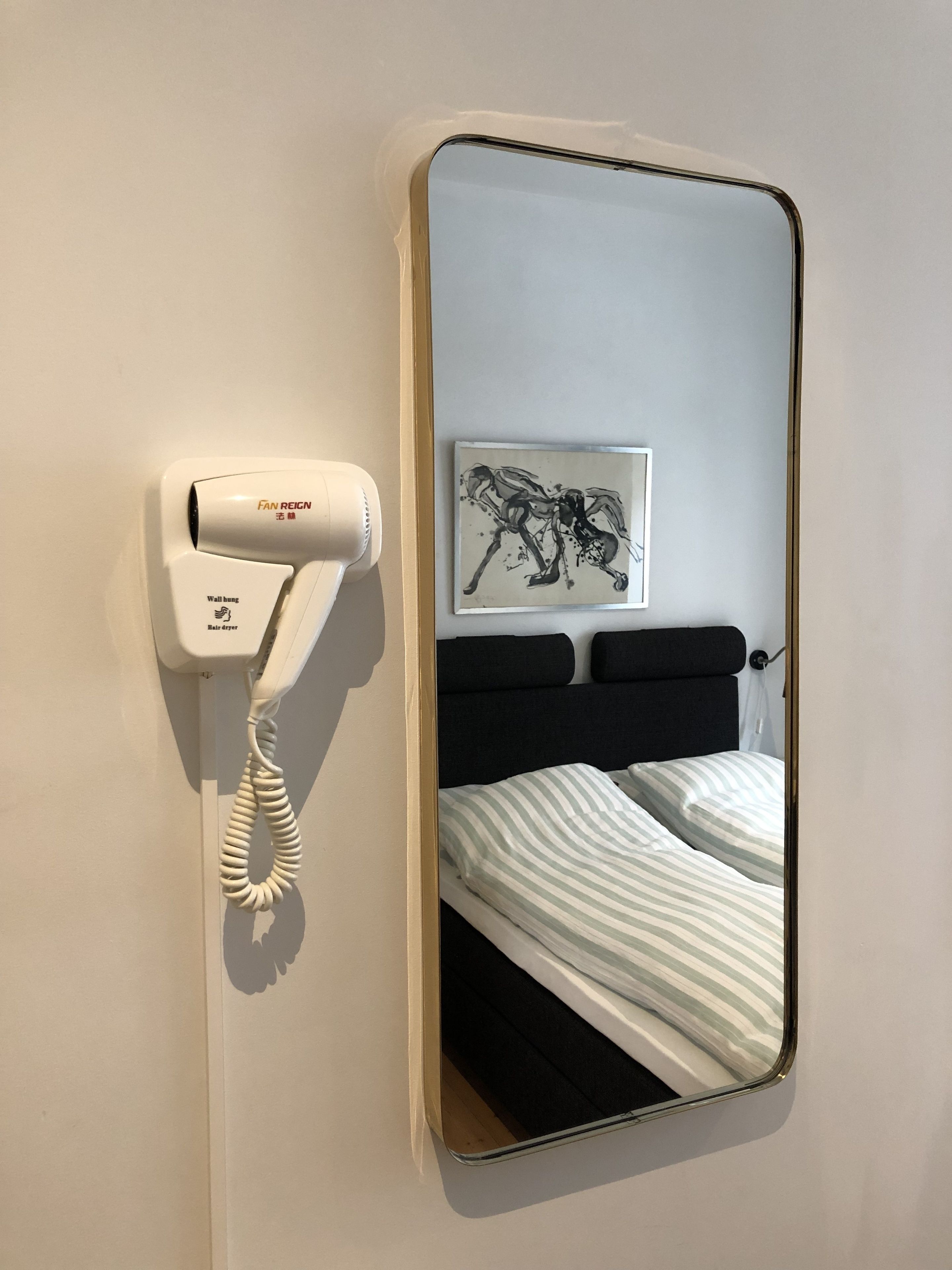 A hotel room with a wall-mounted hair dryer, a framed artwork, and a bed with striped bedding.