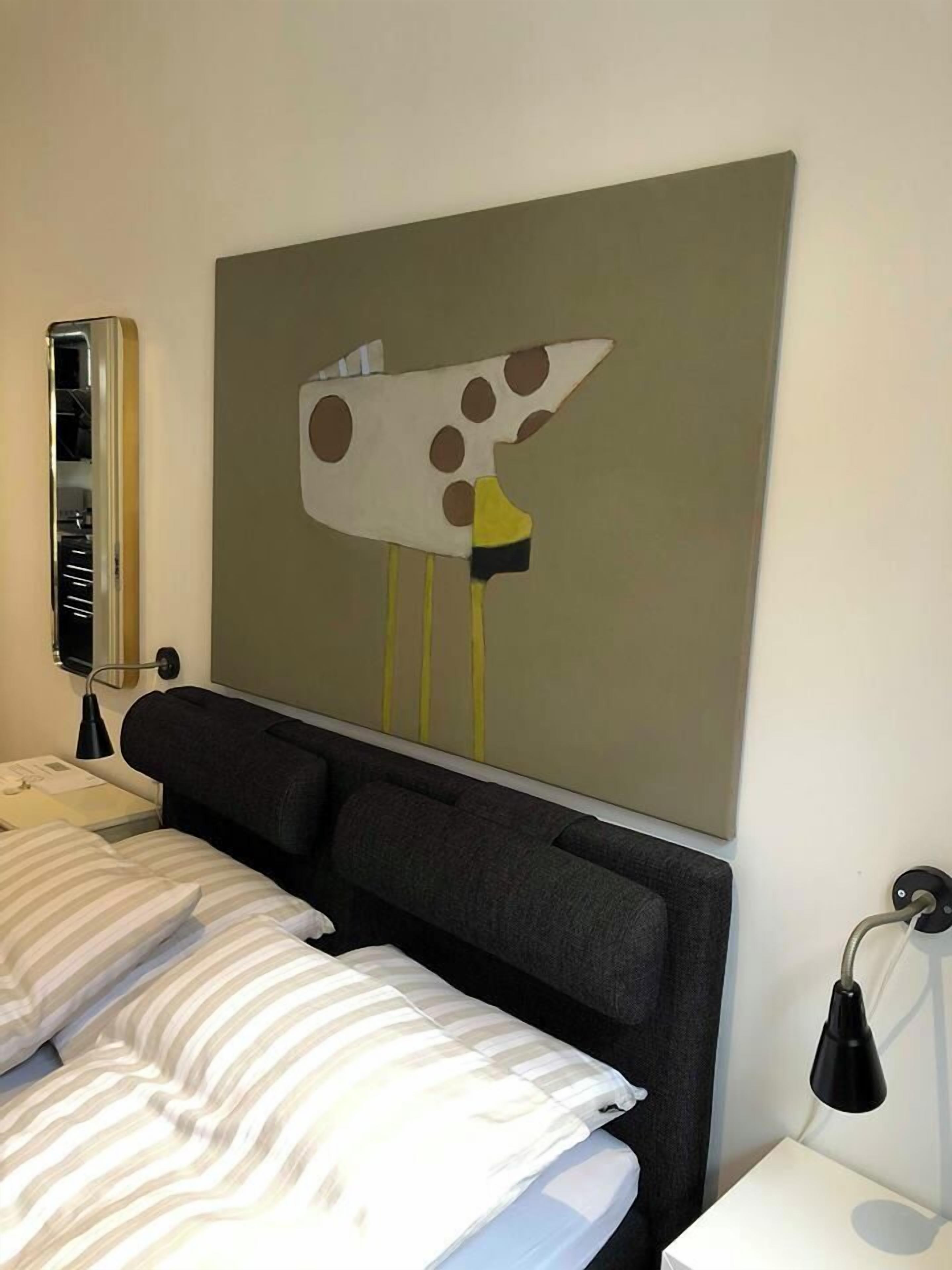 Et hotelværelse med en seng, et natbord med en lampe, og et vægmaleri af et stiliseret dyr.
