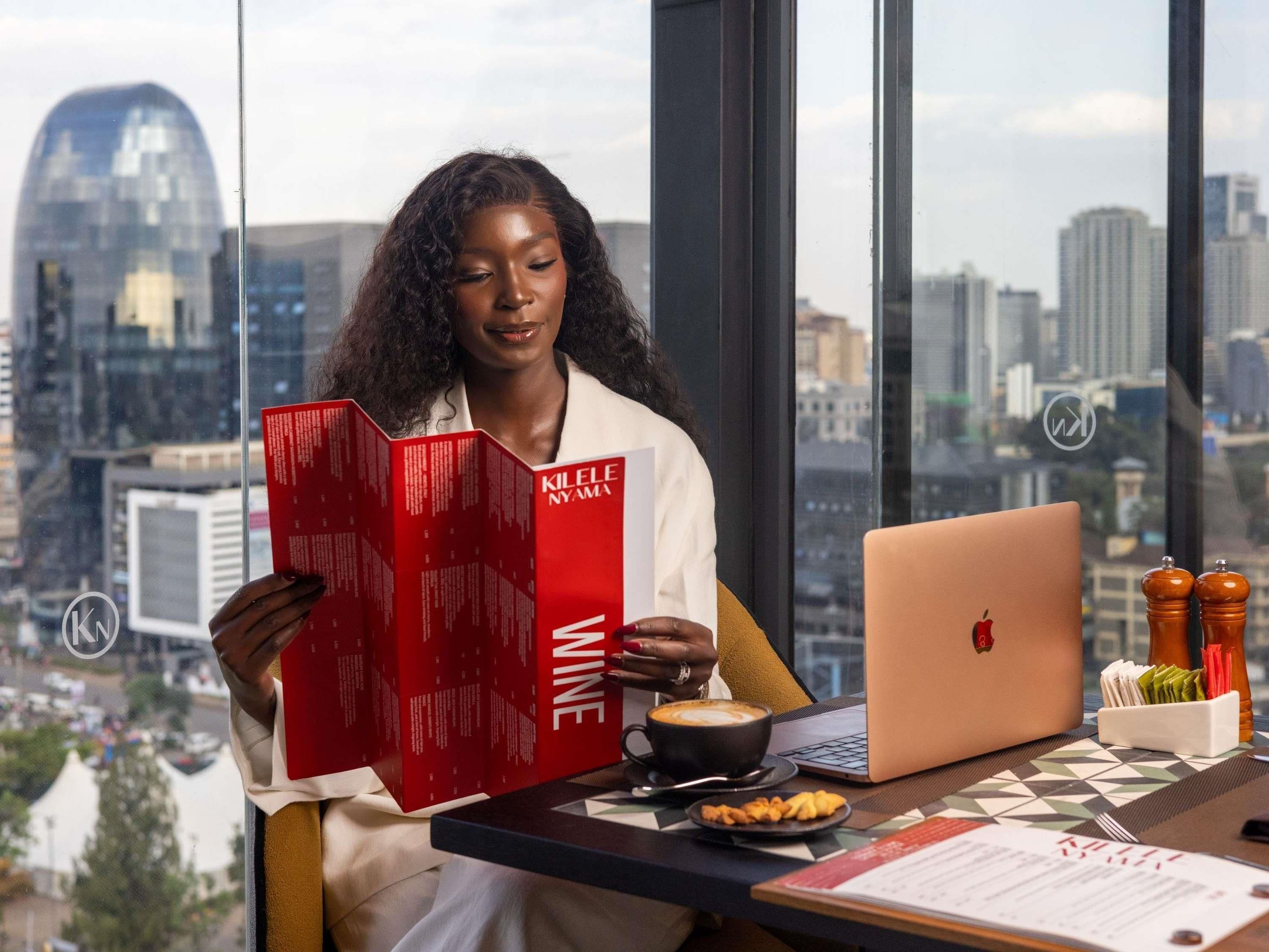 A person reading a red book titled 'KILELE NYAMA' at a desk with a laptop, coffee cup, and a menu.