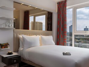 Ein Hotelzimmer mit Bett, Nachttisch mit Kaffeemaschine, Ablagefläche mit Flaschen und Blick auf die Stadt.