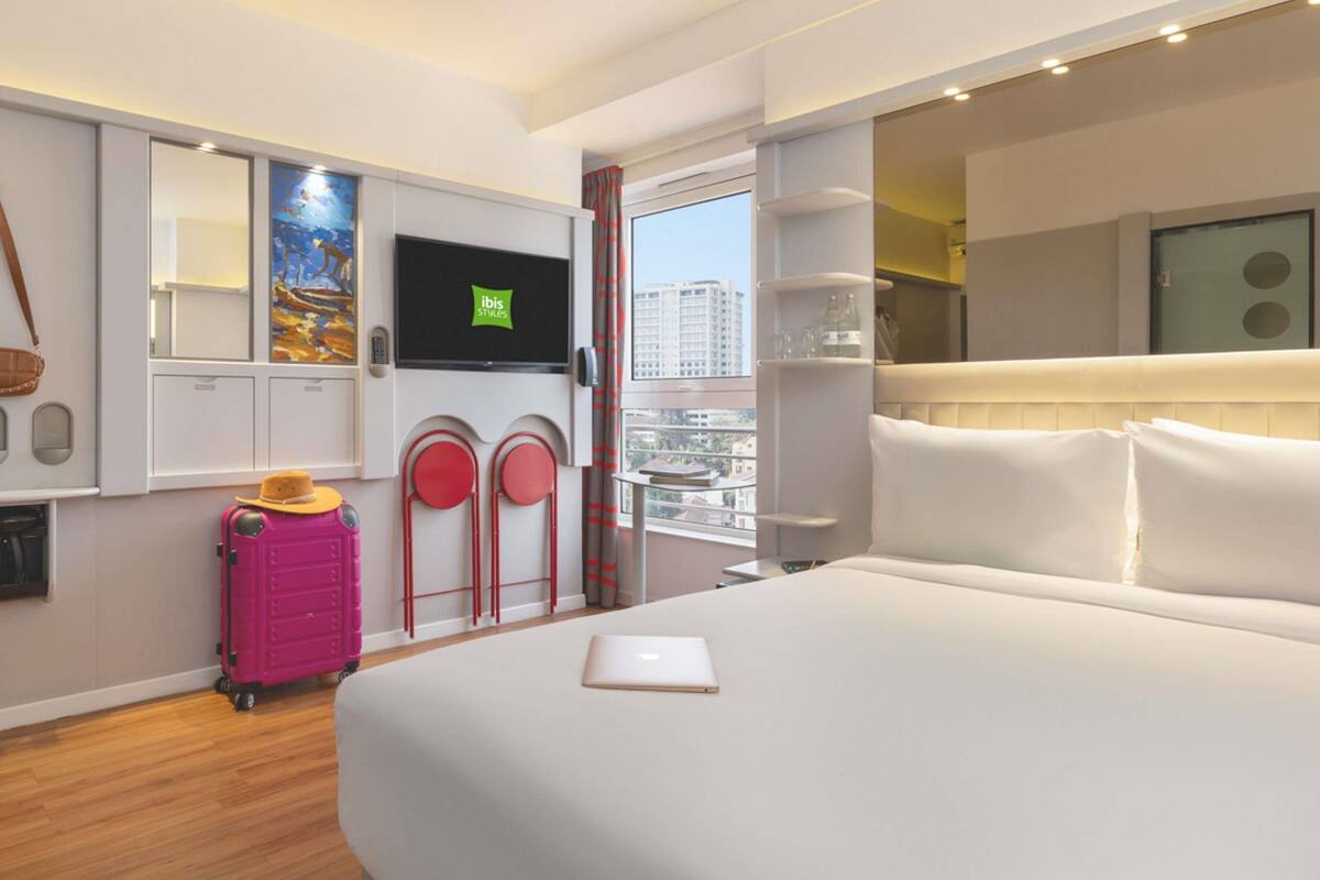 Habitación de hotel con cama, televisión, maleta, un cuadro y vistas a la ciudad.