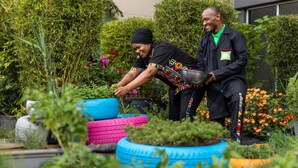 Dos personas trabajando en un jardín exuberante, con mucha vegetación y coloridos maceteros hechos con neumáticos.