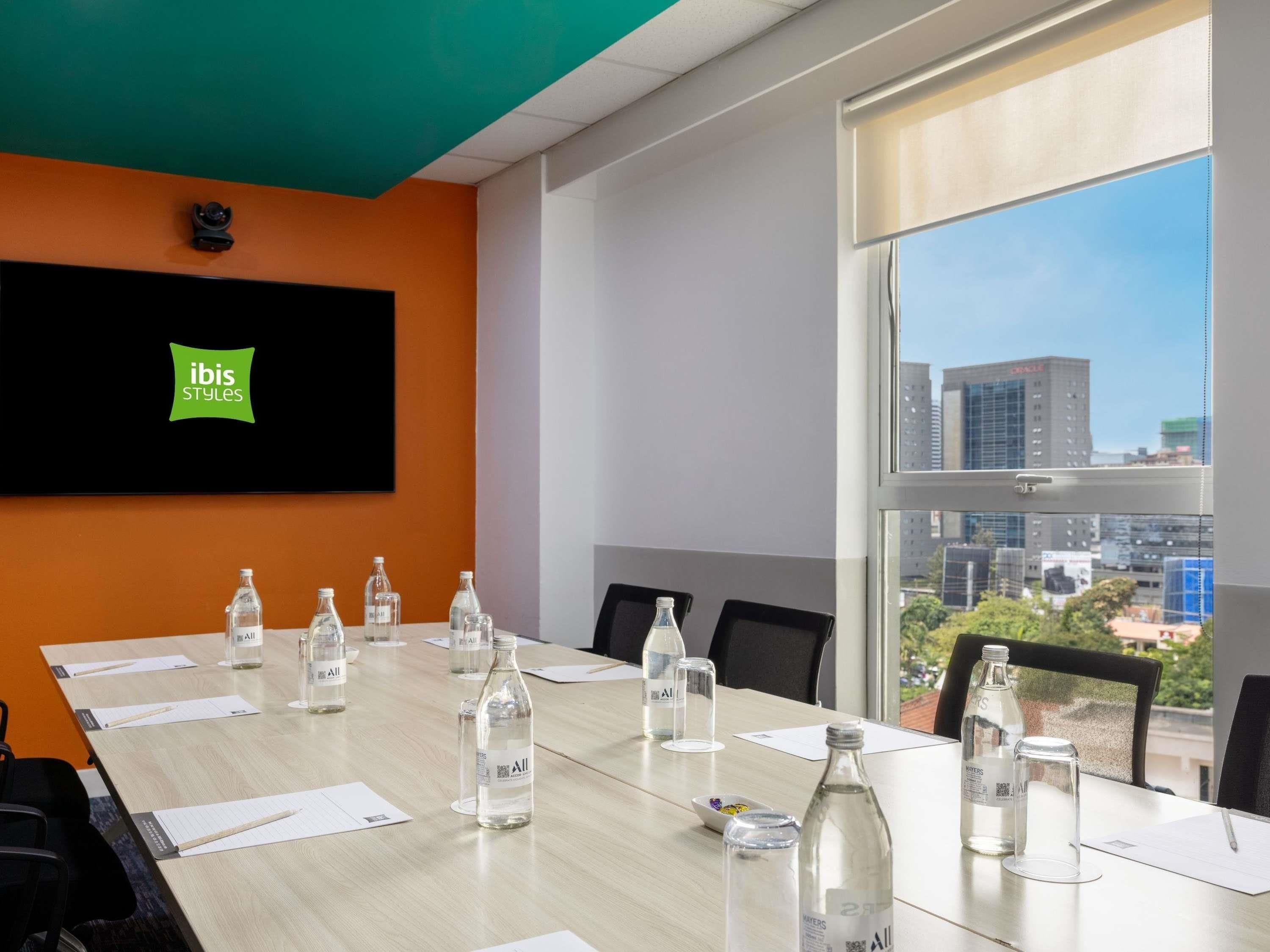 Sala de conferências com uma grande mesa, cadeiras, garrafas de água e vista para a cidade através de uma ampla janela.