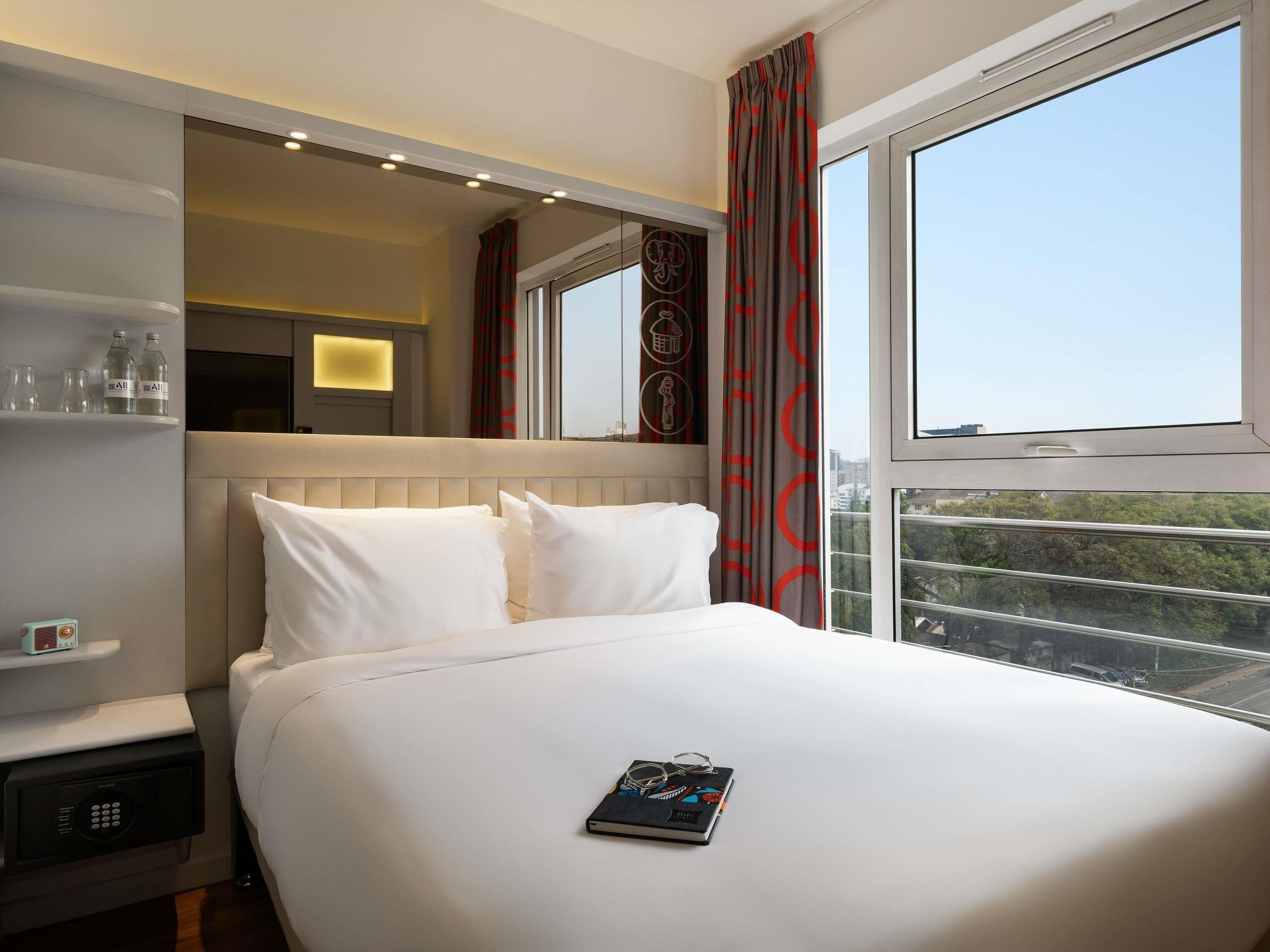 A hotel room with a large bed, a bedside table with a remote control, a mirror, and a window with a city view.