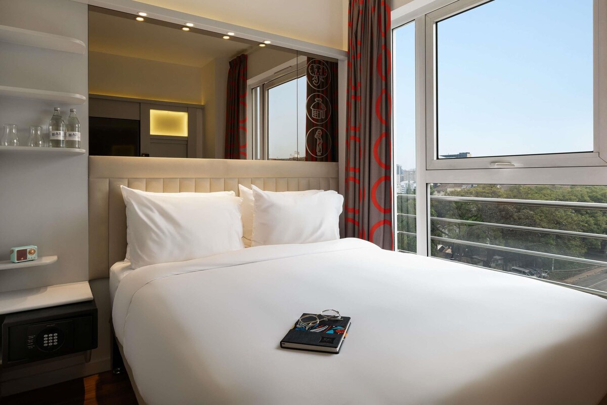 Habitación de hotel con una cama grande, una mesita de noche con control remoto, un espejo y una ventana con vista a la ciudad.