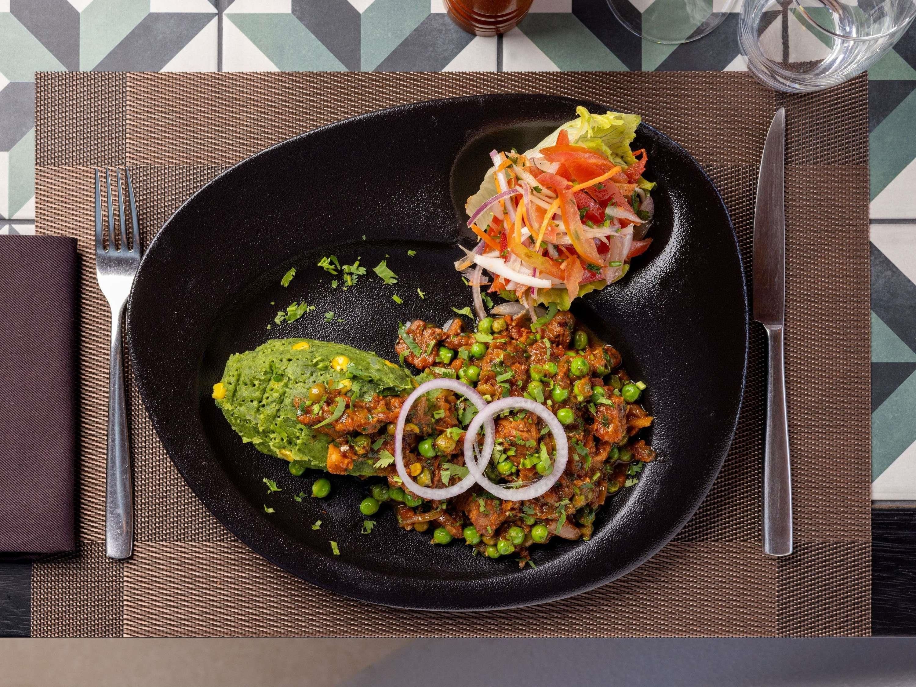Um prato preto com uma refeição que inclui um vegetal verde, um prato de carne com ervilhas e uma salada.
