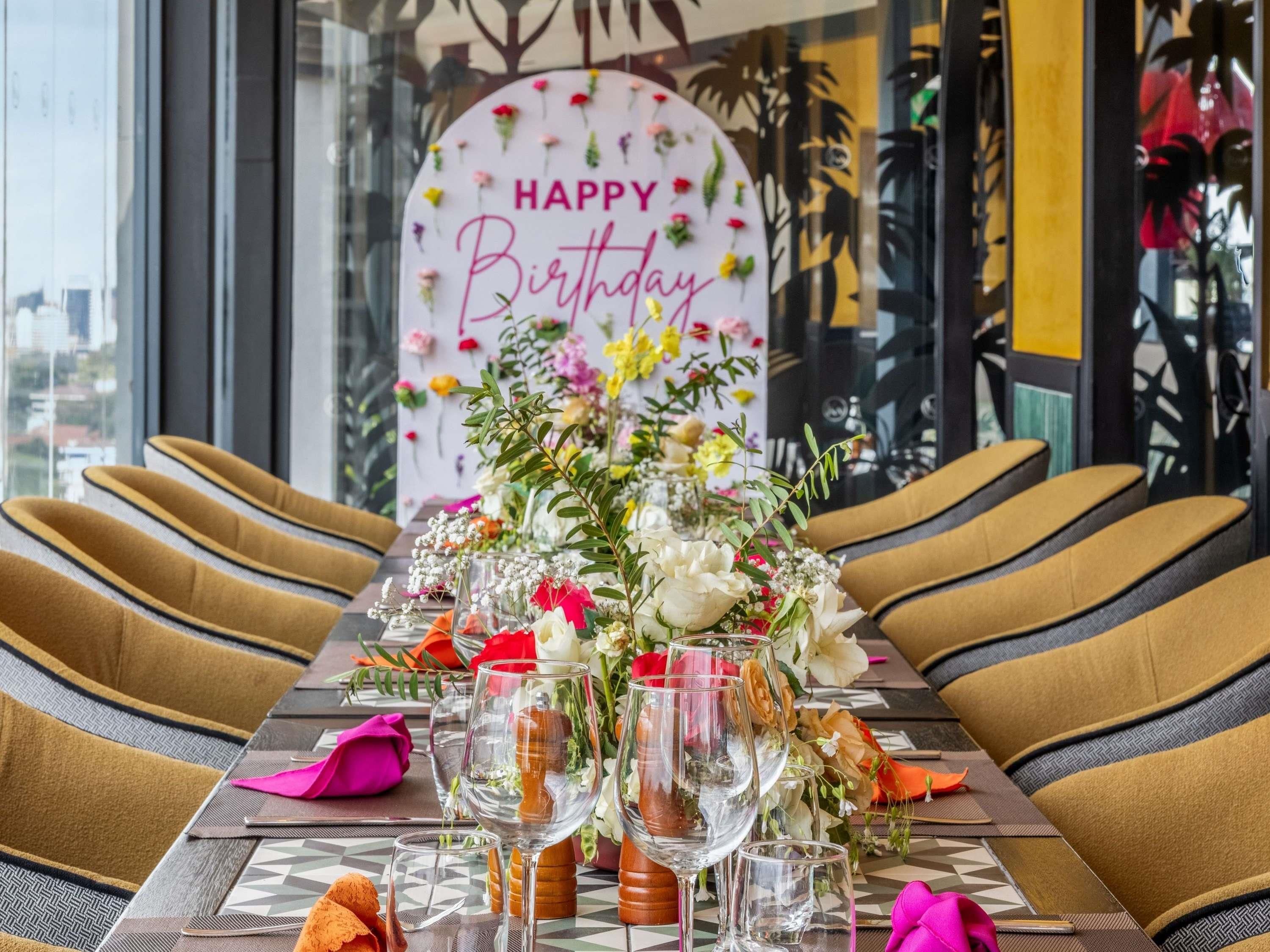 Una mesa de comedor lista con sillas, un centro de mesa floral y un cartel que dice 'Feliz Cumpleaños'.
