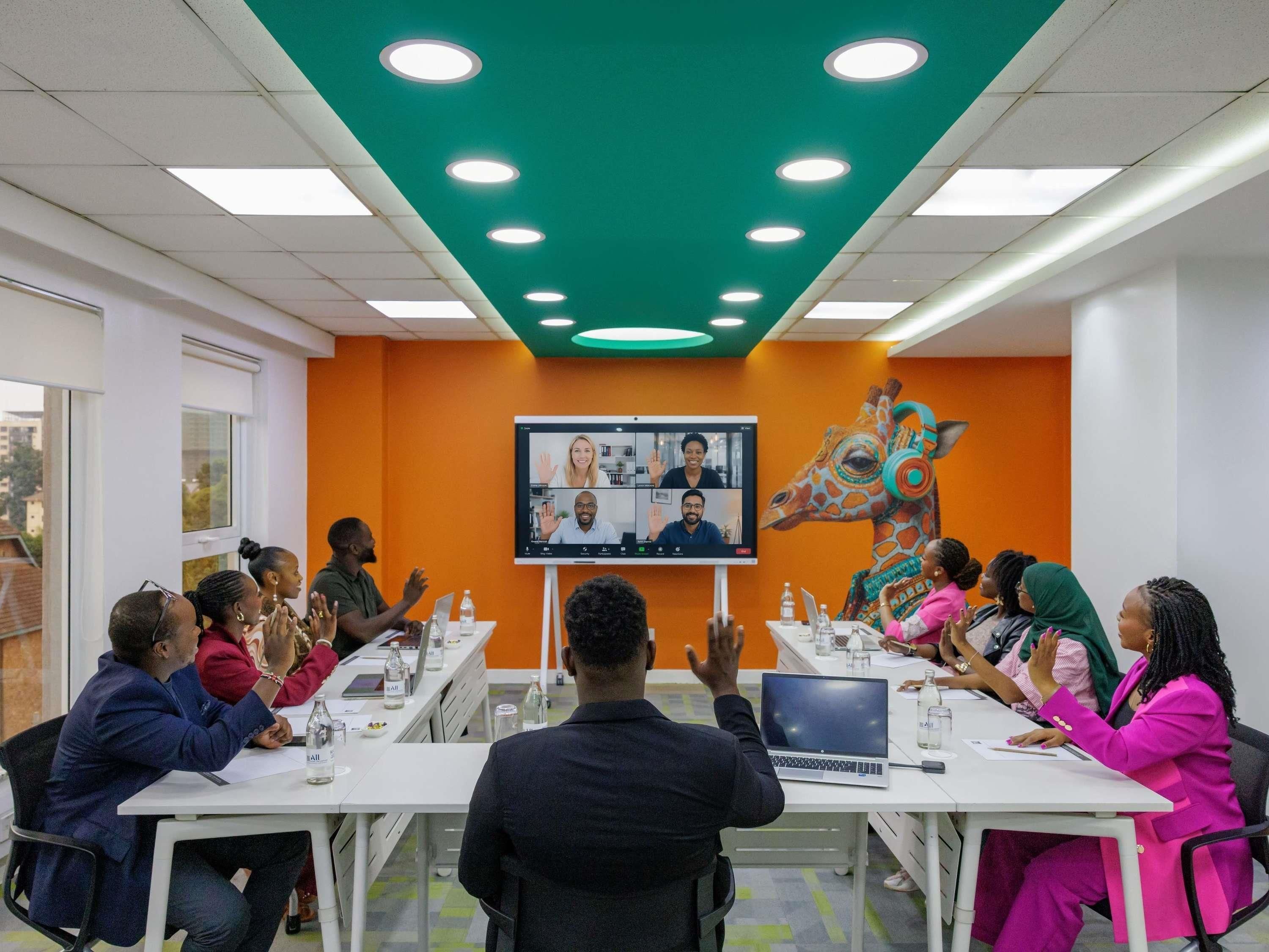 A meeting room with a large screen displaying a video conference, white tables, and chairs arranged for participants.