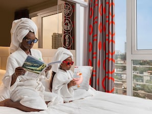 Dos personas vestidas de blanco sentadas en una cama leyendo libros, con vistas a la ciudad.