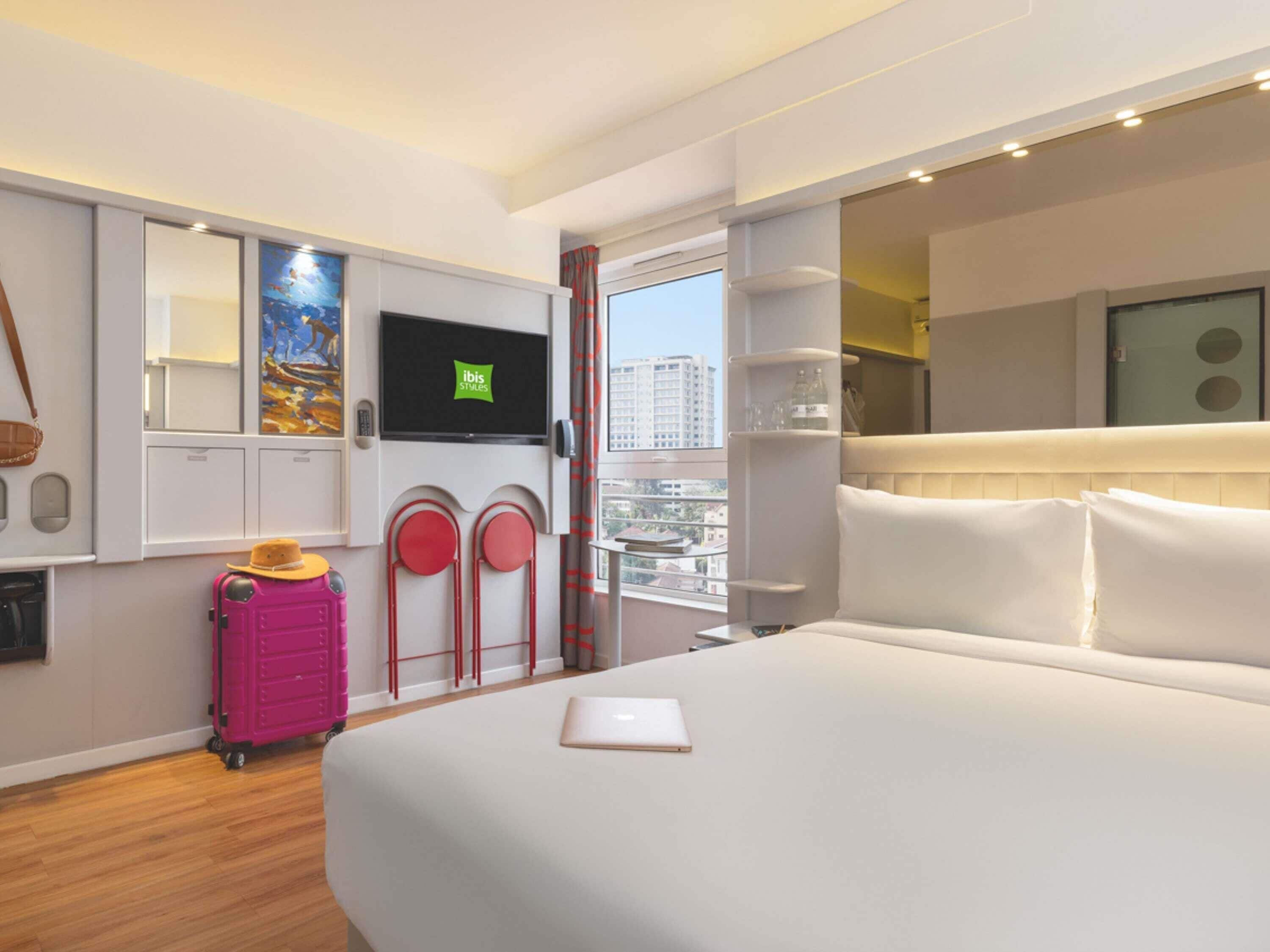 A hotel room with a bed, a television, a suitcase, a painting, and a city view.