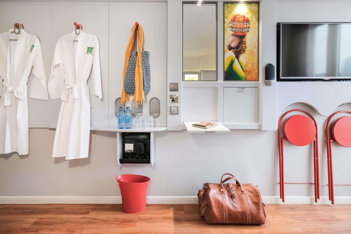 A hotel room with a bed, a red bucket, a brown bag, and a wall-mounted TV.