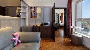 Una habitación de hotel moderna con sofá, una otomana pequeña, zona de comedor con microondas y un ventanal con vistas a la ciudad.