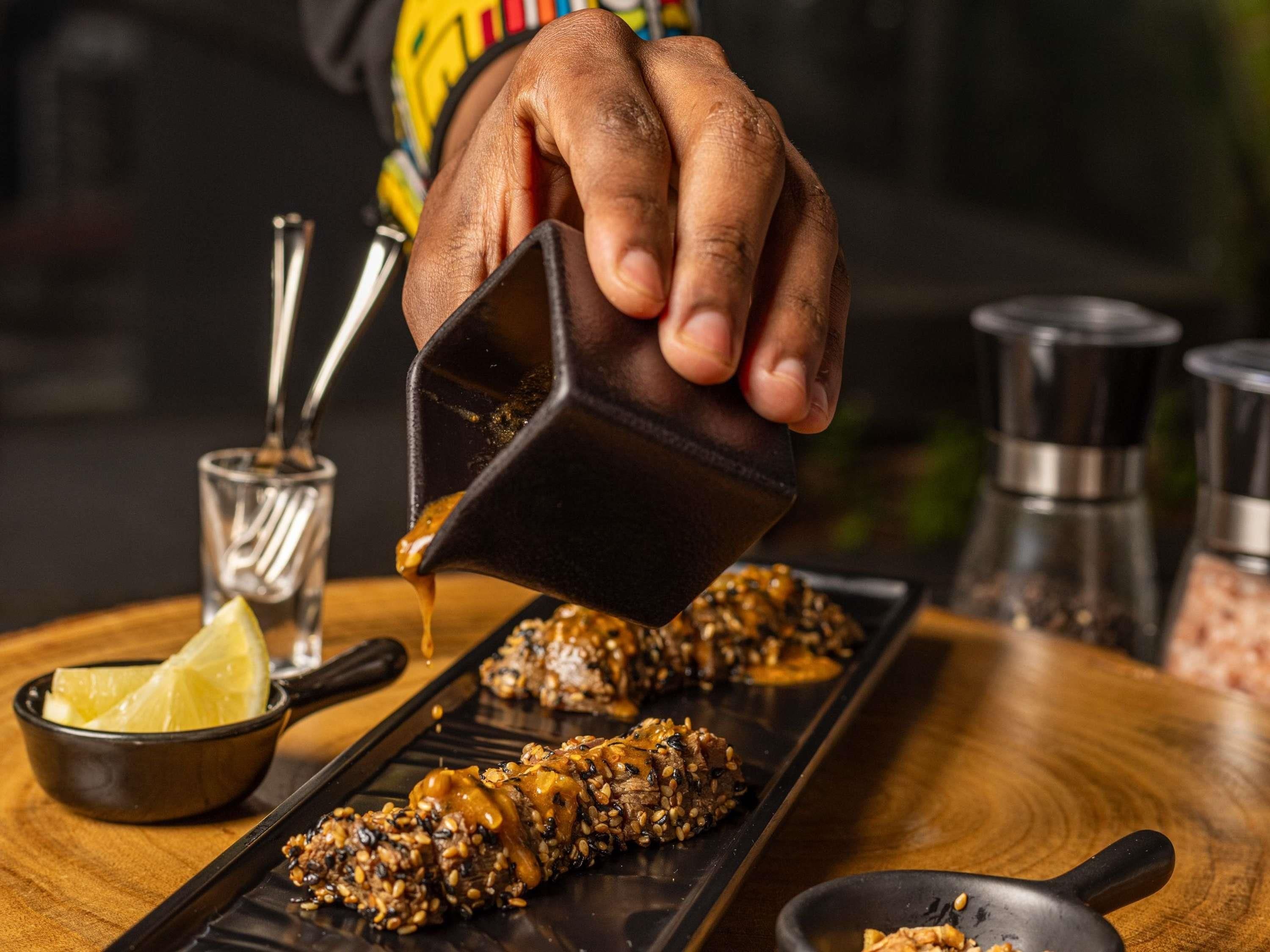 A hand pouring sauce over a dish with sesame seeds.