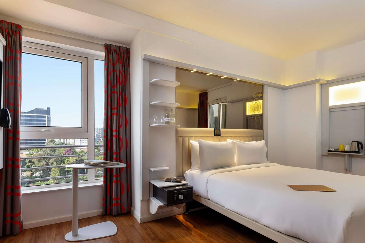 Habitación de hotel con una cama grande, mesitas de noche, una pequeña mesa redonda, microondas y vista a la ciudad por la ventana.
