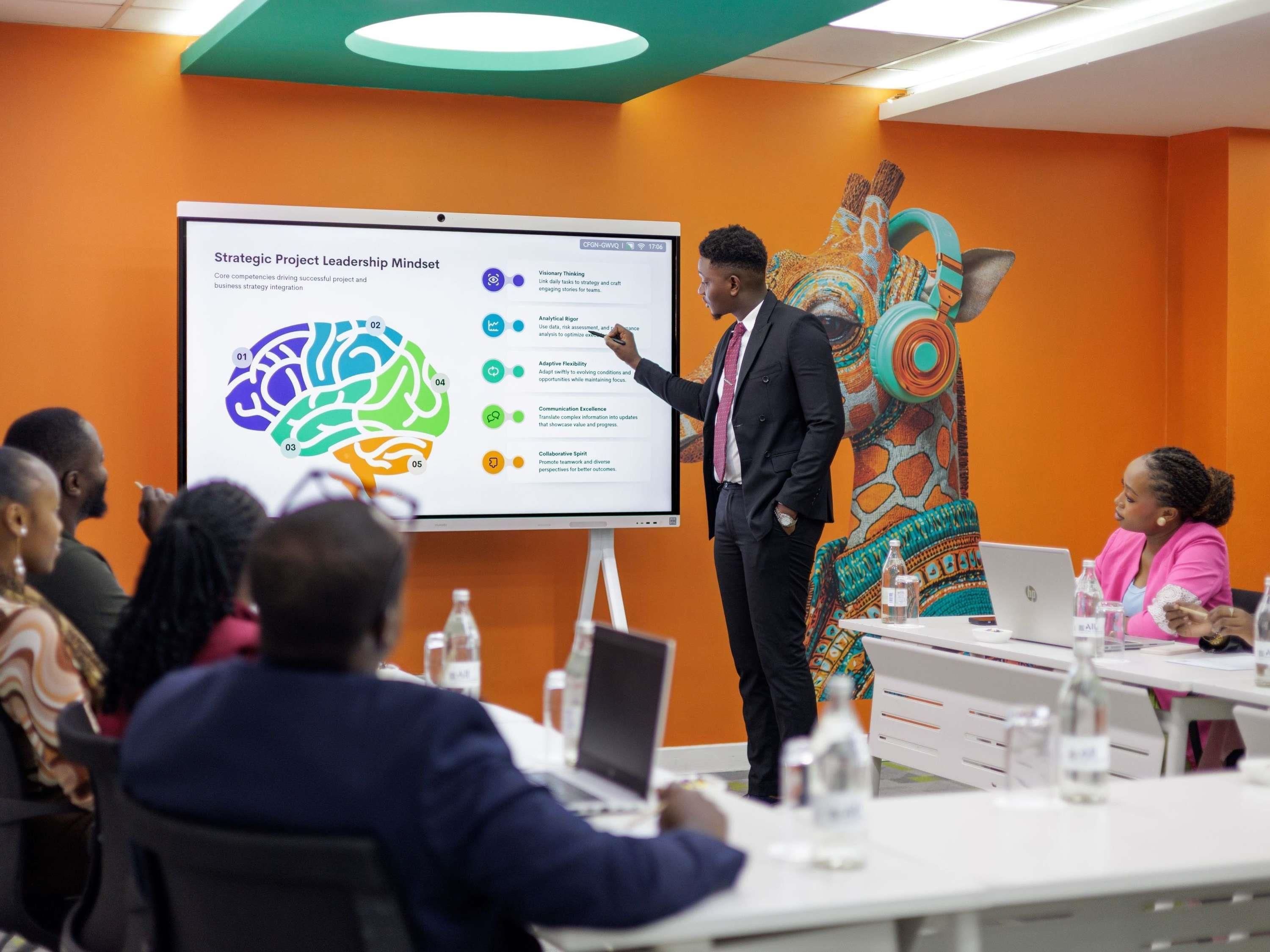 A conference room with a presentation on a screen, a person presenting, and attendees seated at tables with laptops.