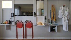 Une chambre d’hôtel moderne équipée d’un téléviseur à écran plat, d’un miroir, d’un fauteuil rouge, d’une petite étagère sur laquelle repose un chapeau, et d’un peignoir accroché au mur.
